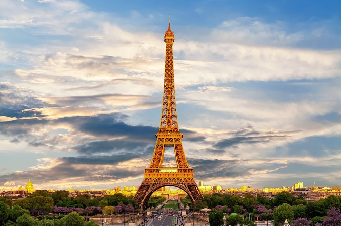 A imagem mostra a Torre Eiffel, um ícone arquitetônico em Paris, França. A torre de ferro aparece ao centro, destacando-se contra um céu parcialmente nublado com tons de azul e dourado, tipicamente ao entardecer. Ao redor da torre, é possível ver algumas árvores e edifícios na base, compondo a paisagem urbana da cidade.
