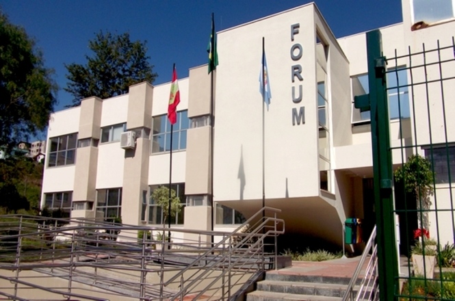 A imagem mostra a fachada de um edifício identificado como "FÓRUM". O prédio tem uma arquitetura moderna com linhas retas e janelas grandes. Há três bandeiras hasteadas na frente, e a entrada é acessível por uma rampa. O dia está claro e ensolarado, sugerindo um ambiente agradável ao redor do edifício.