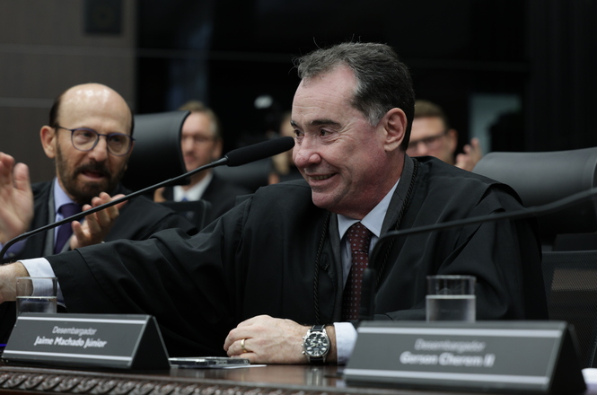 A imagem mostra um homem em um ambiente formal, aparentemente em um tribunal ou sala de conferência. Ele está sorrindo, vestindo um manto preto, típico de magistrados ou oficiais da justiça, e está sentado em frente a um microfone, sugerindo que está participando de uma sessão oficial. Ao seu lado, há outro homem, também vestindo traje formal, que parece estar aplaudindo. Em frente a eles há placas com nomes, indicando posições ou títulos oficiais. O ambiente é sério e profissional.