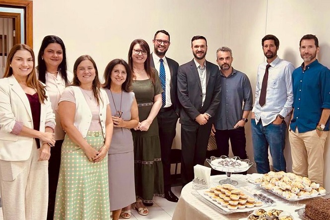 A imagem mostra um grupo de dez pessoas sorrindo para a câmera em um ambiente interno. Elas estão em pé, lado a lado, vestindo trajes formais e casuais. À direita, há uma mesa com diversos alimentos, como doces e salgados. Parece ser uma reunião ou evento social em um ambiente corporativo ou similar.