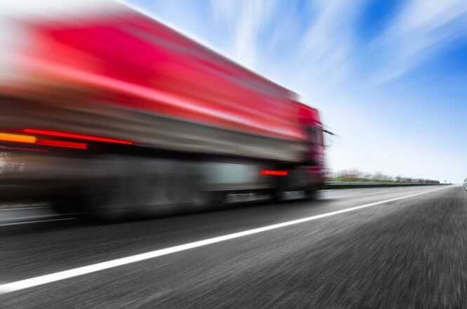 A imagem mostra um caminhão vermelho em movimento rápido em uma estrada. A foto está desfocada, destacando a velocidade do veículo. O céu está azul com algumas nuvens, e a estrada tem marcações brancas. A paisagem ao fundo está igualmente desfocada.