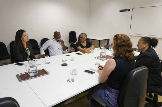A imagem mostra seis pessoas sentadas ao redor de uma mesa de reunião em uma sala. Na mesa, há copos, um jarro de água e itens como celulares e papéis. As pessoas parecem estar em uma discussão ou reunião. O ambiente é formal, com cadeiras pretas e uma parede clara ao fundo, onde também há um quadro branco.