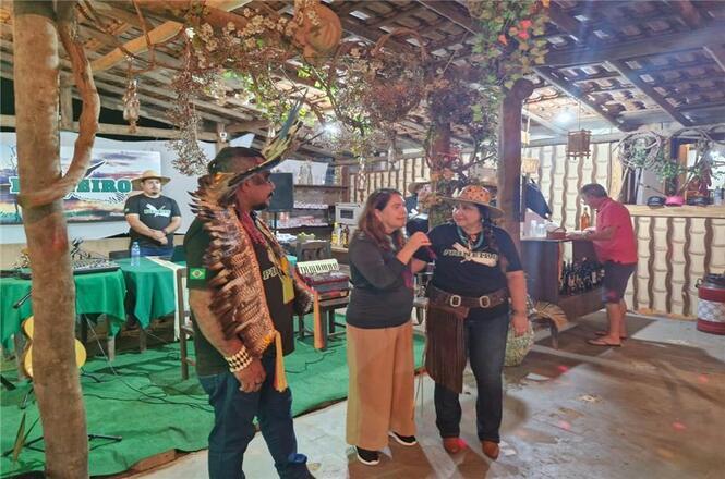 A imagem mostra um ambiente interno decorado de forma rústica, com três pessoas conversando no centro. Duas delas estão vestidas com trajes típicos regionais, incluindo chapéus e adereços. No fundo, há mesas com coberturas verdes, objetos decorativos e outro homem próximo ao que parece ser um balcão. A iluminação do local é suave, com luzes penduradas no teto.