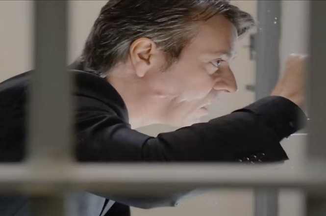 A imagem mostra um homem de perfil, vestindo um terno escuro. Ele parece estar olhando ou examinando algo através de janelas ou grades. A expressão facial sugere concentração ou análise. O enfoque está no lado direito do rosto e no gesto da mão direita. A cena é iluminada, destacando o cabelo e as feições do homem.