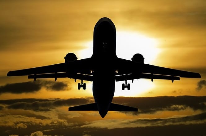 A imagem mostra a silhueta de um avião voando contra um céu ao pôr do sol. O sol está parcialmente coberto por nuvens, criando uma iluminação dramática por trás da aeronave. A cena tem um tom alaranjado e dourado, típico do final da tarde.