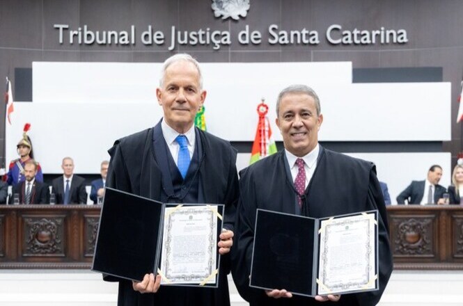 A imagem mostra duas pessoas usando trajes formais, segurando documentos em frente a uma bancada. Ao fundo, há uma placa com a inscrição "Tribunal de Justiça de Santa Catarina". Atrás deles, há outras pessoas sentadas em roupas formais. A cena parece ser uma cerimônia oficial.