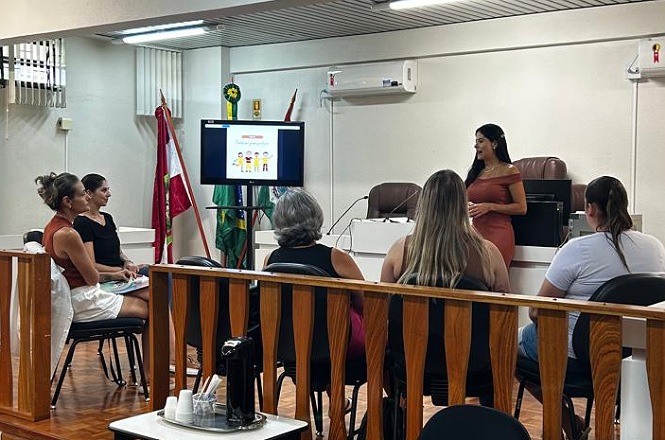 A imagem mostra um ambiente interno onde uma apresentação está acontecendo. Uma mulher está em pé, apresentando algo para um grupo de pessoas sentadas em cadeiras à sua frente. Ela está usando um vestido laranja. Na frente do grupo, há um monitor exibindo o que parece ser uma apresentação de slides. Há bandeiras ao fundo e algumas mobílias como uma mesa grande e cadeiras. O ambiente parece ser um auditório ou sala de conferências. Uma cafeteira com copos está sobre uma mesa à frente.