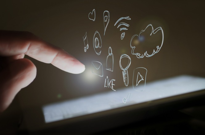 A imagem mostra um dedo humano apontando para uma tela iluminada, que provavelmente é de um dispositivo eletrônico como um tablet ou smartphone. Sobre a tela, há ícones desenhados flutuando no ar, representando várias funções e conceitos digitais, como um envelope (mensagem), um coração, uma nota musical, um símbolo de Wi-Fi, uma nuvem e outros elementos relacionados à comunicação e mídias sociais. O fundo é escuro e destaca a luz emanando da tela.