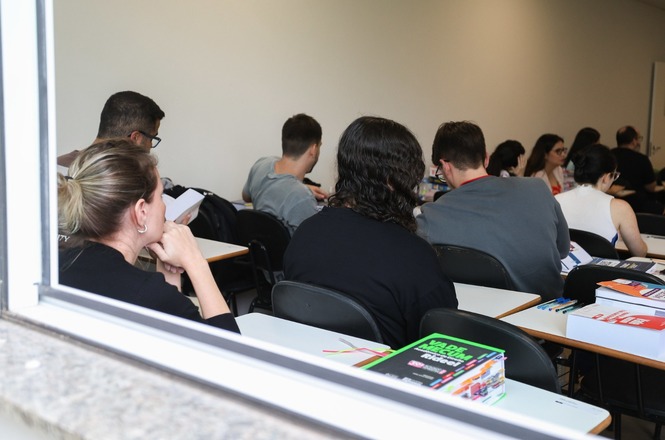 A imagem mostra uma sala de aula vista de fora, através de uma janela. Há várias pessoas sentadas em carteiras, aparentemente estudantes, algumas lendo e outras escrevendo. Na mesa, há livros e materiais escolares. O ambiente tem uma iluminação clara e as pessoas estão voltadas para frente, como se estivessem assistindo a uma aula ou uma apresentação.