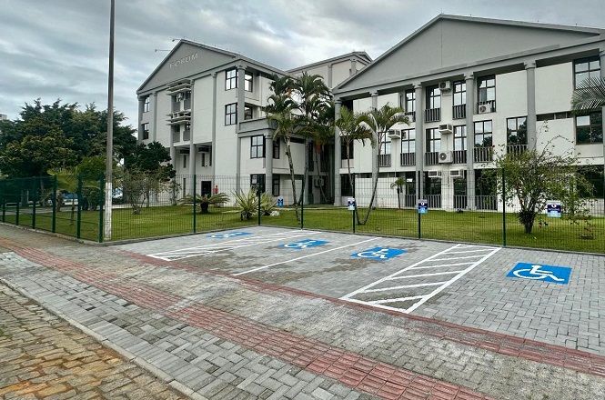 A imagem mostra um prédio com arquitetura moderna, identificado como "FÓRUM". Há uma área de estacionamento com vagas para deficientes, marcadas com o símbolo de acessibilidade azul no chão. A área ao redor é cercada por gramado e algumas palmeiras. O céu está nublado, sugerindo um clima possivelmente chuvoso ou de tempo fechado.