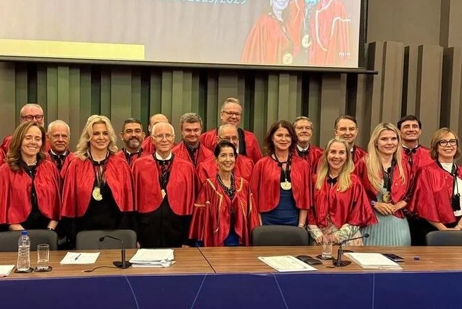 A imagem mostra um grupo de pessoas reunidas, vestindo becas vermelhas com medalhas penduradas no pescoço. Eles estão em um ambiente interno, possivelmente uma cerimônia ou evento formal, como uma graduação ou evento acadêmico. Há uma mesa à frente com papéis, microfones, uma garrafa de água, e ao fundo, uma tela de projeção. Todos estão posando para uma foto conjunta.