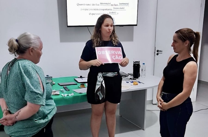 Na imagem, há três pessoas em pé participando de uma atividade em um ambiente interno. A pessoa no centro está segurando um cartaz que diz "OBSERVAÇÃO". Ao fundo, há uma mesa com vários objetos e um projetor exibindo texto em uma tela. Parece ser um ambiente de aprendizado ou reunião.
