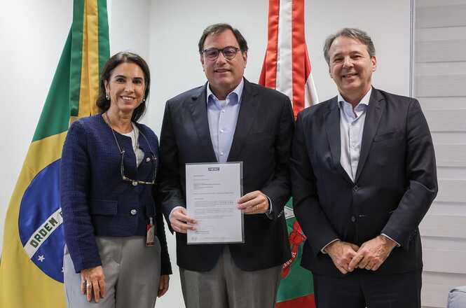 Foto horizontal: Francisco Oliveira Neto segura documento assinado entre autoridades e bandeiras do Brasil e SC.