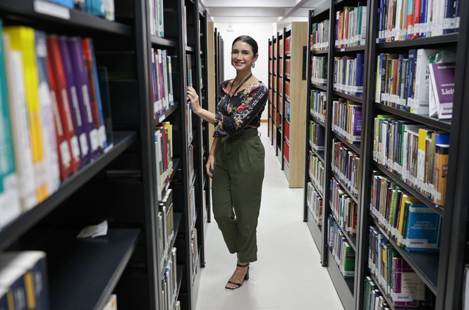 A imagem mostra uma mulher em pé entre prateleiras de livros em uma biblioteca. Ela está sorrindo e usando uma blusa estampada de manga longa e calças verde-oliva. As prateleiras estão repletas de livros de várias cores e tamanhos. O ambiente é bem iluminado e organizado, com corredores largos permitindo fácil acesso aos livros.