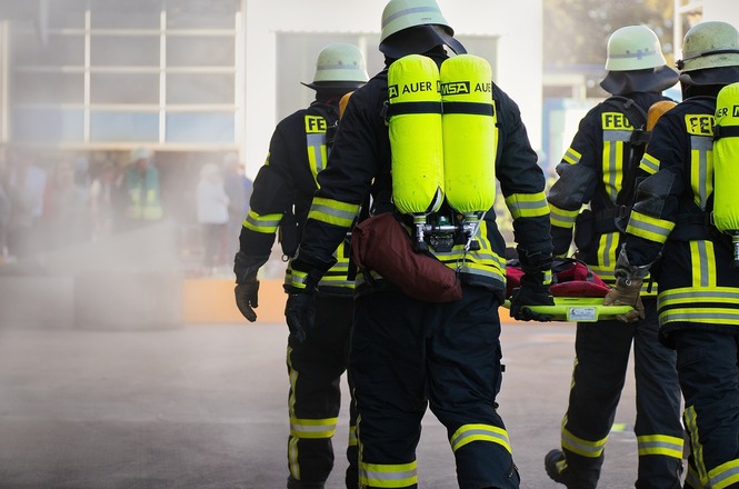 A imagem mostra um grupo de bombeiros caminhando. Eles estão usando equipamentos de proteção completa, incluindo capacetes e máscaras de respiração, e carregam cilindros de ar nas costas. A cena parece estar em um ambiente com fumaça ou vapor, sugerindo que estão em operação ou treinamento. A visão é de trás, destacando os trajes e equipamentos dos bombeiros.