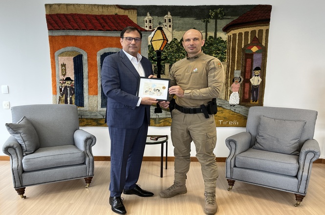 Na imagem, há duas pessoas em pé, parecendo participar de uma cerimônia ou encontro formal. À esquerda, um homem de terno segura uma moldura, enquanto à direita, um indivíduo com uniforme militar ou policial segura a moldura junto com ele. Ao fundo, há uma grande tapeçaria colorida com o desenho de uma cena urbana, incluindo casas e pessoas. Em frente a eles, há duas poltronas cinzas. A cena se passa em um ambiente interno.