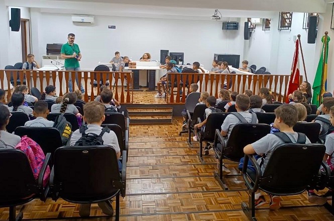 A imagem mostra uma sala de reunião ou tribunal onde várias crianças estão sentadas em cadeiras, voltadas para a frente. Na frente, um adulto em pé está falando. Algumas crianças estão sentadas atrás de uma bancada elevada, que parece ser uma área de tribunal. Há bandeiras em um canto da sala e vários monitores de computador. O piso é de madeira.