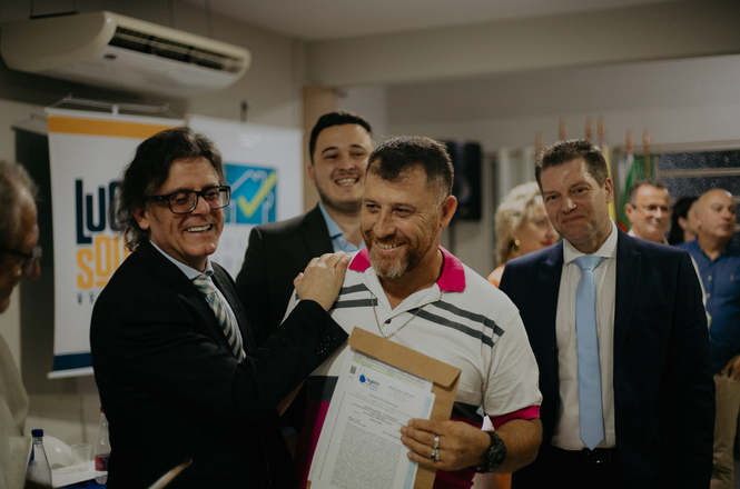 A imagem mostra um grupo de pessoas em um ambiente fechado, provavelmente durante um evento ou cerimônia. Um homem de óculos e terno escuro está ao lado de outro homem que segura um certificado. Ambos estão sorrindo e parecem estar felizes. Ao fundo, há mais pessoas assistindo, algumas delas também vestindo terno. Há um banner visível à esquerda com parte de um logotipo e texto, além de um aparelho de ar-condicionado acima. A atmosfera é de celebração ou reconhecimento.