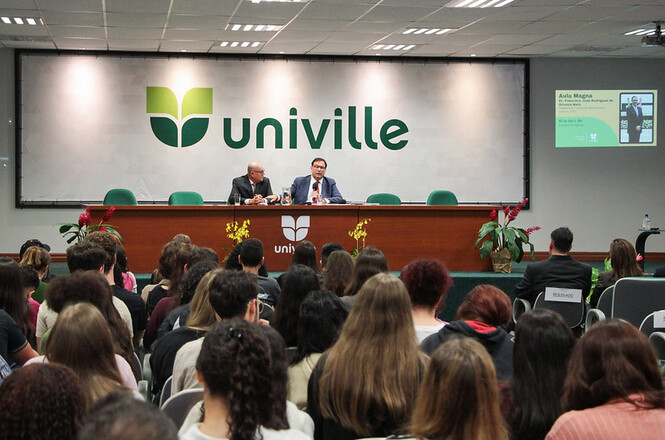 Foto horizontal: Francisco Oliveira Neto fala ao microfone em auditório da Univille, público ao fundo, tons verde e neutros.