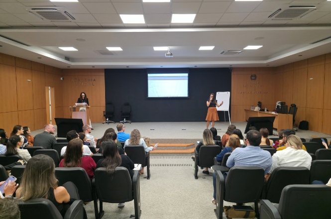 A imagem mostra uma sala de conferências ou auditório, onde várias pessoas estão sentadas, aparentemente assistindo a uma apresentação. Na frente, uma pessoa está em pé falando, enquanto há um projetor exibindo algo em uma tela. Outra pessoa está em posição de apoio, talvez controlando o conteúdo no laptop sobre a mesa. O ambiente é profissional, com paredes de madeira clara, e parece ser uma apresentação empresarial ou educacional.