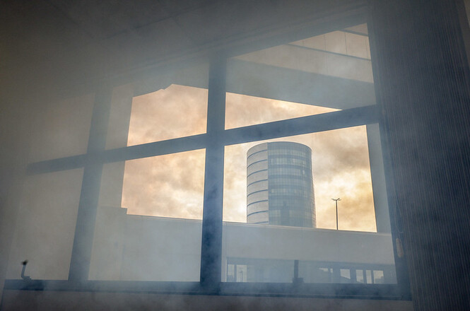 Na imagem, há uma janela vista do interior de um edifício, através da qual se vê o exterior. Fora da janela, um prédio cilíndrico e alto é visível. O céu tem uma aparência enevoada ou embaçada, dando um tom alaranjado à cena. Há também alguma fumaça ou neblina presente dentro ou próximo da janela, que cria uma atmosfera um pouco misteriosa.