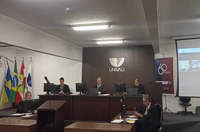 A imagem mostra uma sala de aula ou auditório da UNIVALI, com um ambiente organizado como um tribunal. Há várias pessoas sentadas em uma bancada elevada, possivelmente simulando juízes em um julgamento. Eles estão de frente para computadores sobre a bancada. À esquerda, há bandeiras de estados ou instituições. No canto inferior, um homem está sentado em frente a um computador com documentos, provavelmente representando advogados ou apresentando um caso. Há também um banner à direita com os dizeres "60 Direito UNIVALI". O ambiente sugere uma simulação ou prática educacional de um julgamento.