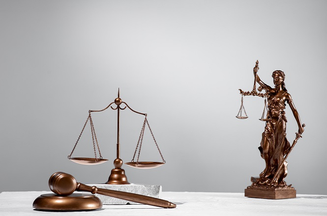 A imagem mostra objetos associados à justiça. À esquerda, há uma balança, símbolo da justiça e da equidade. À frente, vê-se um malhete, usado por juízes para manter a ordem em tribunal. À direita, está uma estátua da deusa Justitia, segurando uma balança e uma espada, representando a imparcialidade e a autoridade da justiça. O fundo é simples, destacando os objetos.