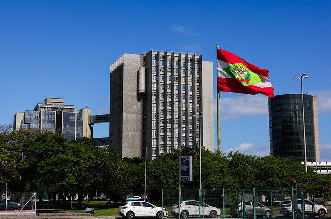 A imagem mostra um complexo de edifícios modernos em um dia ensolarado. No centro, há um prédio alto com arquitetura sólida e linhas retas. À direita, uma bandeira grande está hasteada e tremulando ao vento. À esquerda, nota-se um outro edifício com design contemporâneo, e à direita, um prédio cilíndrico de vidro. Na frente dos prédios, há árvores e carros estacionados. O céu está azul com poucas nuvens.