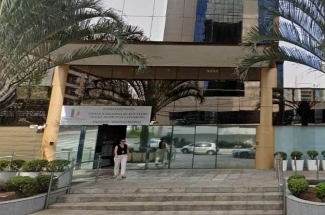 A imagem mostra a entrada de um edifício comercial com fachada de vidro. Há uma escada de acesso com corrimão e algumas plantas decorativas ao redor. Na entrada, está uma placa que parece indicar o nome ou a função do prédio, mencionando uma unidade regional de negócios. Algumas palmeiras e arbustos estão dispostas ao redor da área. Uma pessoa está na frente do prédio, próxima à entrada.
