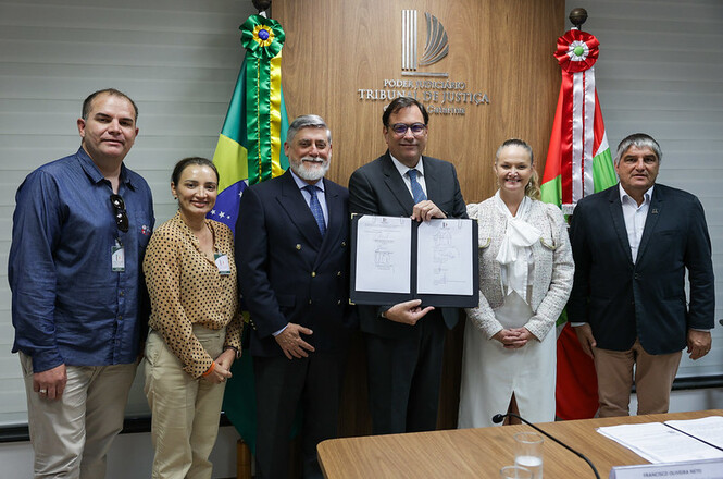 Na imagem, há seis pessoas posando para uma foto em frente a uma parede com o emblema do "Poder Judiciário Tribunal de Justiça de Santa Catarina". Cinco delas estão ao redor de um homem que segura um documento em um quadro. Todos estão vestidos formalmente. Há bandeiras do Brasil e de Santa Catarina ao fundo. Parece ser um evento oficial ou assinatura de documentos.