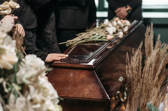 A imagem mostra um caixão de madeira cercado de flores e arranjos florais. Uma mão repousa sobre o caixão, sugerindo um momento de despedida ou homenagem. Há pessoas ao redor vestindo roupas escuras, típicas de um funeral. O ambiente é solene e respeitoso.