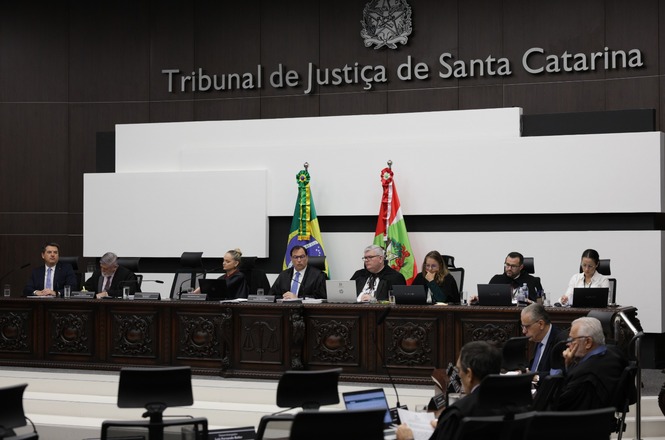 A imagem mostra uma sessão em um tribunal, especificamente o Tribunal de Justiça de Santa Catarina. Várias pessoas estão sentadas em uma mesa, possivelmente juízes ou autoridades jurídicas, todos usando ternos formais. Ao fundo, veem-se as bandeiras do Brasil e de Santa Catarina, o que indica a localização e a jurisdição do tribunal. Existem computadores e documentos sobre a mesa, sugerindo que se trata de uma reunião ou julgamento. O ambiente é formal, com um design que reflete a seriedade e a autoridade de um tribunal de justiça.