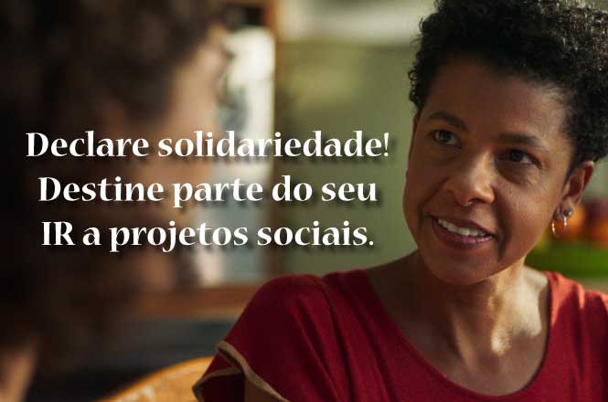 A imagem mostra uma mulher sorrindo e conversando com outra pessoa que está fora de foco. Ao lado, há um texto dizendo: "Declare solidariedade! Destine parte do seu IR a projetos sociais." O fundo está desfocado, dando ênfase à mensagem e às pessoas na cena.