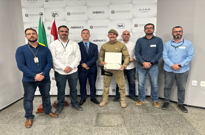 A imagem mostra um grupo de sete homens posando para uma foto em um ambiente fechado. Ao fundo, há um banner com logotipos e duas bandeiras em suportes. O homem ao centro está vestindo um uniforme militar e segurando uma placa ou troféu. Os outros homens estão vestidos de forma casual ou formal, com camisas e ternos. O ambiente parece ser uma sala de conferências ou um local semelhante, com piso de granito.
