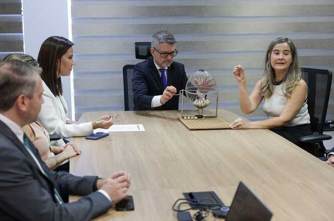 Na imagem, há um grupo de seis pessoas em torno de uma mesa de madeira em um escritório. No centro da mesa, há um globo de sorteio com uma pessoa manuseando-o, sugerindo que eles estão participando de um sorteio ou seleção aleatória. As pessoas estão vestidas formalmente, e na mesa há papéis, um laptop e um celular. A sala possui iluminação natural entrando por uma janela.