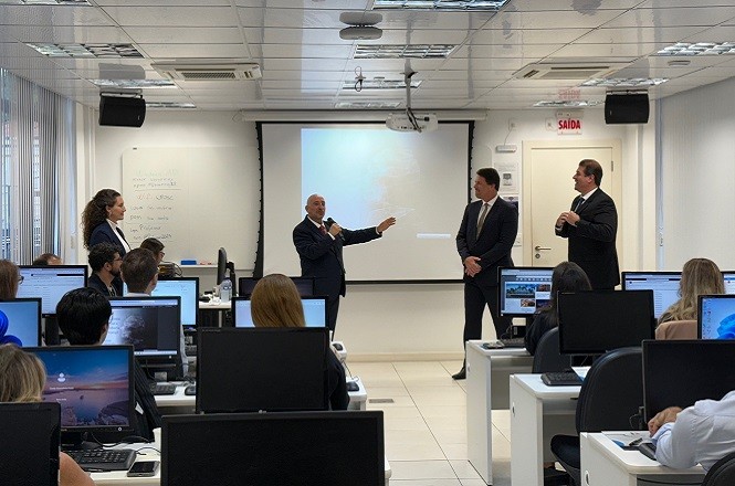 A imagem mostra uma sala de aula ou treinamento com várias pessoas sentadas em frente a computadores. Na frente da sala, três homens de terno estão em pé, parecendo participar de uma apresentação ou discussão. Um deles está falando ao microfone, enquanto os outros dois estão escutando. Existe uma tela de projeção ao fundo, mas não está claramente visível o que está projetado. A sala parece moderna, com iluminação brilhante e equipamentos tecnológicos.