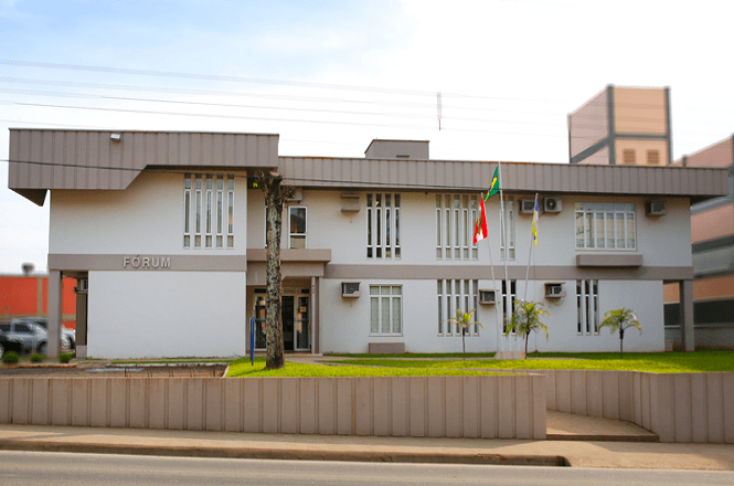 A imagem mostra um edifício de dois andares com a palavra "FÓRUM" na fachada. Ele tem um design moderno com janelas grandes e quadradas. Na frente, há um gramado bem cuidado com três mastros de bandeira, dois dos quais têm bandeiras hasteadas. O prédio está rodeado por uma cerca baixa, e ao lado, há uma rua pavimentada.