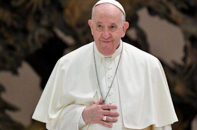 A imagem mostra um homem idoso vestido com trajes religiosos, incluindo uma veste branca e um solidéu. O fundo da imagem está desfocado. Ele parece estar em um ambiente interno e está olhando na direção da câmera.