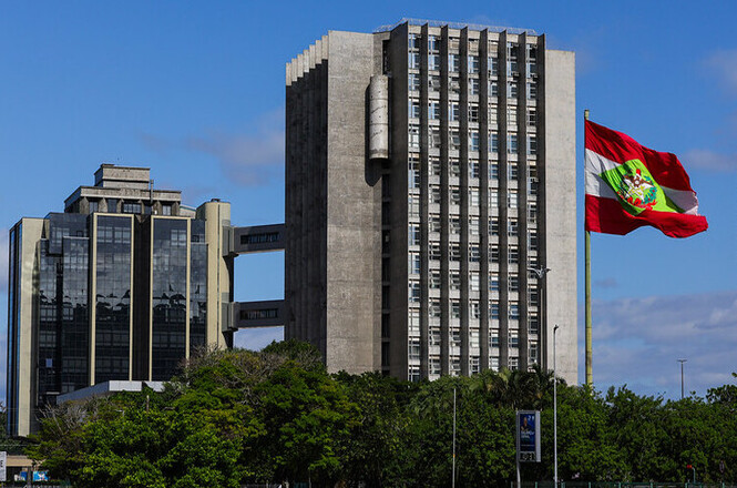 A imagem mostra dois prédios altos em um cenário urbano. À esquerda, há um edifício com janelas espelhadas e uma estrutura moderna. À direita, existe um prédio mais simples com linhas verticais e uma aparência de concreto. Em frente aos prédios, há uma grande bandeira colorida hasteada, destacando-se contra o céu azul ao fundo. A área ao redor dos edifícios tem vegetação abundante com árvores verdes.