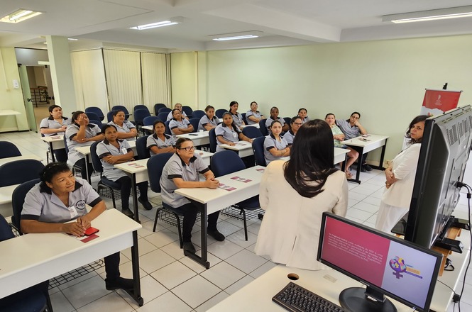 A imagem mostra uma sala de aula com várias pessoas sentadas em fileiras, prestando atenção ao que parece ser uma apresentação. Todos estão vestindo uniformes semelhantes. Há uma mulher de pé na frente da sala, de costas para a câmera, apresentando algo em um monitor. A sala tem mesas brancas e cadeiras azuis, e há materiais de papel sobre as mesas. O ambiente é iluminado e parece ser um espaço para treinamento ou aula.
