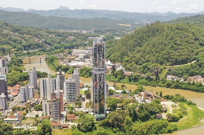 A imagem mostra uma cidade rodeada por áreas verdes e montanhas ao fundo. No centro, há um edifício alto e esbelto que se destaca entre as construções mais baixas ao redor. A cidade parece estar situada próximo a um rio, que atravessa o cenário. Há uma ponte visível cruzando o rio e conectando diferentes partes da cidade. A vegetação é exuberante, com muitas árvores, e o cenário parece ser em uma região montanhosa.