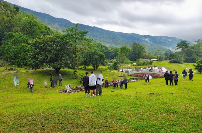 A imagem mostra um grupo de pessoas em um campo verde, localizado em uma área cercada por árvores e montanhas ao fundo. Há um pequeno lago ou rio à direita, com uma ponte sobre ele. O céu está nublado, proporcionando uma atmosfera um pouco cinzenta. As pessoas parecem estar passeando ou participando de uma atividade ao ar livre.