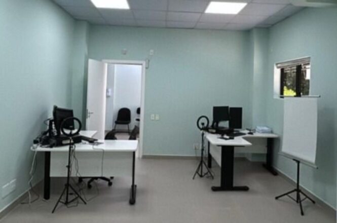 A imagem mostra um escritório com duas mesas brancas equipadas com computadores e monitores. Há cadeiras de escritório, e cada mesa possui um suporte para microfone. Na parede direita, há uma janela. À direita, também está presente um quadro branco em um suporte. No fundo, uma porta aberta revela outra sala com uma cadeira. As paredes são pintadas em um tom claro de verde-azulado.