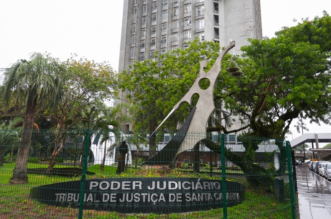 A imagem mostra o prédio do Tribunal de Justiça de Santa Catarina, com uma escultura moderna na frente. A escultura tem um design abstrato e está cercada por um jardim com algumas árvores e plantas. Na frente, há uma placa escrita "Poder Judiciário, Tribunal de Justiça de Santa Catarina". O edifício ao fundo é alto e tem um estilo arquitetônico robusto. A cena também parece estar em um dia chuvoso ou nublado, pois o chão está molhado.
