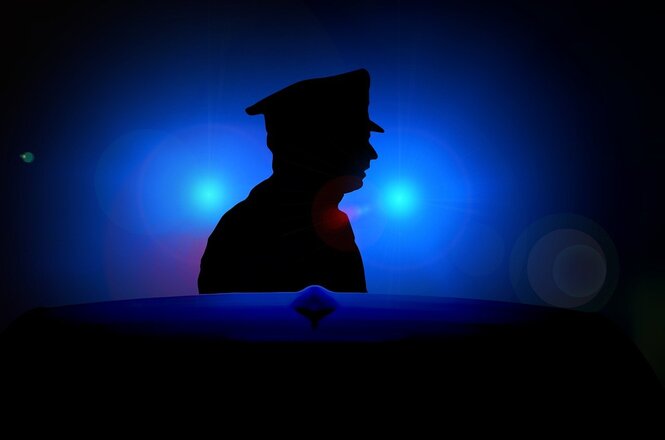 A imagem mostra a silhueta de uma pessoa usando um chapéu, que parece ser um uniforme de policial, contra um fundo azul iluminado por luzes brilhantes. A silhueta está no centro, vista de perfil, e é realçada por um forte contraste entre o escuro da figura e o brilho das luzes ao fundo. A atmosfera é de mistério ou autoridade.