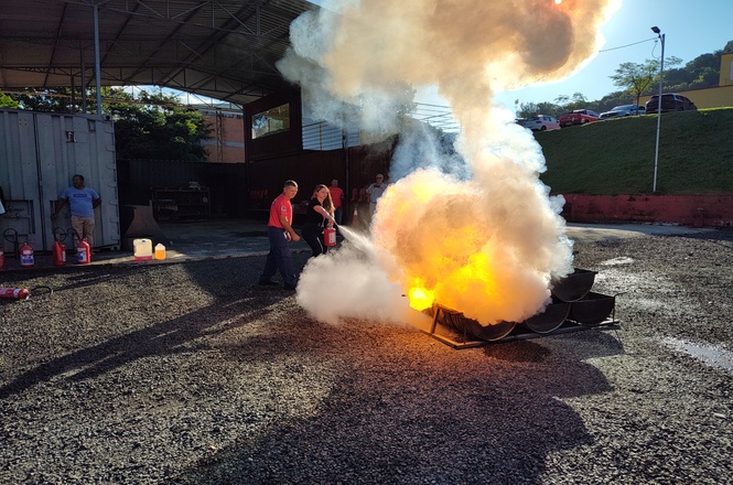 A imagem mostra algumas pessoas participando de um treinamento de combate a incêndio. Há uma chama intensa e muita fumaça saindo de um recipiente no chão. As pessoas estão usando extintores de incêndio para apagar o fogo. Parece ser uma simulação ao ar livre, com um local coberto ao fundo onde há alguns equipamentos de incêndio e materiais de suporte. O ambiente é cercado por uma área gramada e veículos no estacionamento.