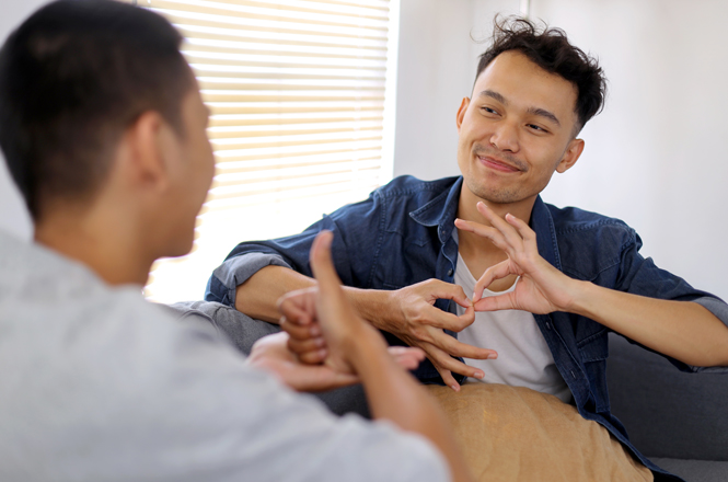 A imagem mostra duas pessoas sentadas em um ambiente fechado. Uma delas está virada de costas para a câmera e a outra está de frente, com uma expressão de sorriso, aparentemente engajadas em uma conversa. A pessoa que está de frente está usando linguagem de sinais, formando gestos com as mãos. O ambiente é iluminado por luz natural que entra por uma janela com persianas. O cenário parece ser descontraído e amigável.