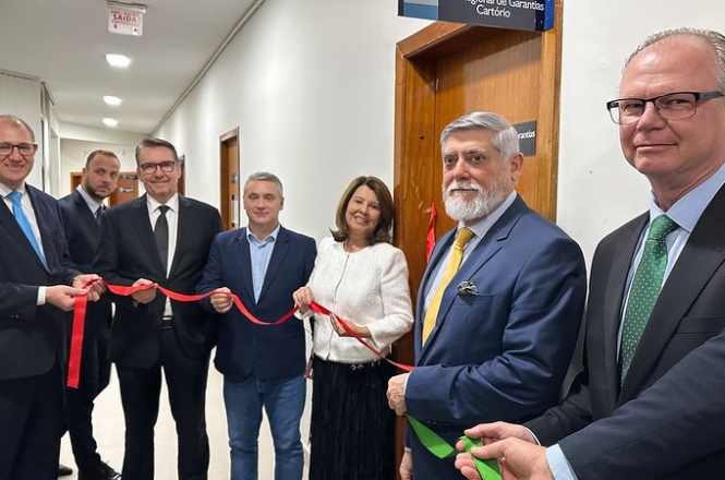 Na imagem, várias pessoas estão participando de uma cerimônia de inauguração ou abertura, possivelmente em um corredor de um edifício. Elas estão segurando uma fita vermelha, simbolizando a inauguração de um espaço. Todos estão vestidos formalmente, indicando a importância do evento. Há um letreiro acima de uma porta ao fundo, sugerindo a entrada para um local específico. As expressões são de expectativa e satisfação.