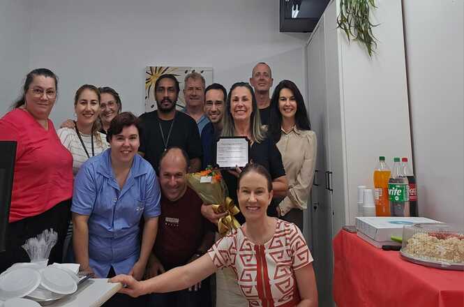 A imagem mostra um grupo de pessoas em um ambiente fechado, possivelmente em uma sala de escritório ou de confraternização. Elas estão posando para a foto, algumas sorrindo, e parece que estão comemorando uma ocasião especial. Há uma mesa à direita com bebidas em garrafas e algumas travessas, que podem conter alimentos. Uma das pessoas está segurando um buquê de flores e outra segura o que parece ser uma pequena placa ou um certificado.