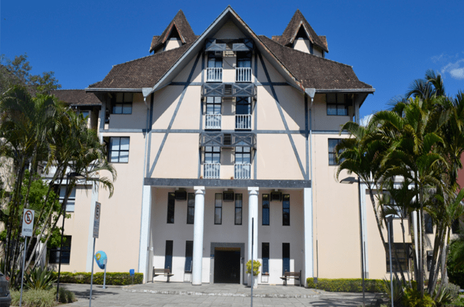 A imagem mostra um edifício com arquitetura de estilo enxaimel. Ele possui um telhado inclinado de cor escura e várias janelas distribuídas de maneira simétrica. Na frente do edifício, há colunas brancas e uma entrada central. Ao redor, há árvores e arbustos, com um céu azul ao fundo.